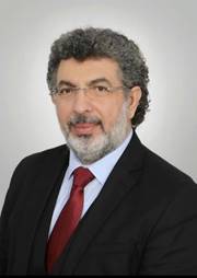 The image shows a man wearing a dark suit and red tie, with a distinctive curly hair texture.AI-generated content may be incorrect.